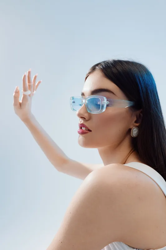 Young woman with long dark hair wearing transparent sunglasses and white top poses with raised arm on light background. Concept of beauty fashion and modern style portrait.