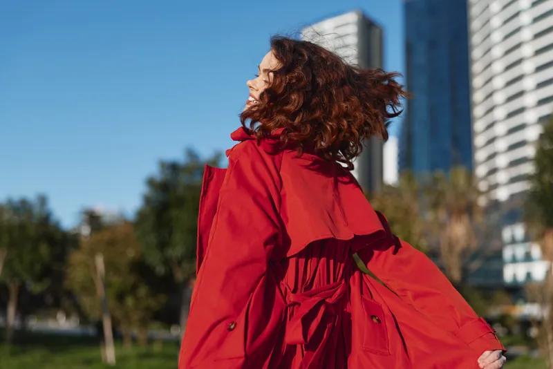 Confident woman in a bright red coat stands in an urban park as wind blows her hair, turning away from the camera, showcasing bold street style in a windy city setting, with tall buildings and clear