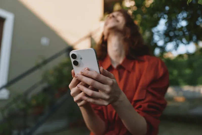 Woman lifestyle outdoor shot holding smartphone looking up with relaxed mood, casual red blouse, sunlit background, candid moment, contemporary selfie