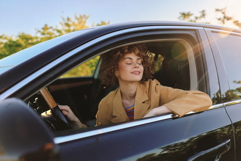 A young woman with curly hair drives a car, eyes closed, smiling, immersed in a sunny moment with a retro film color vibe and a sense of freedom.