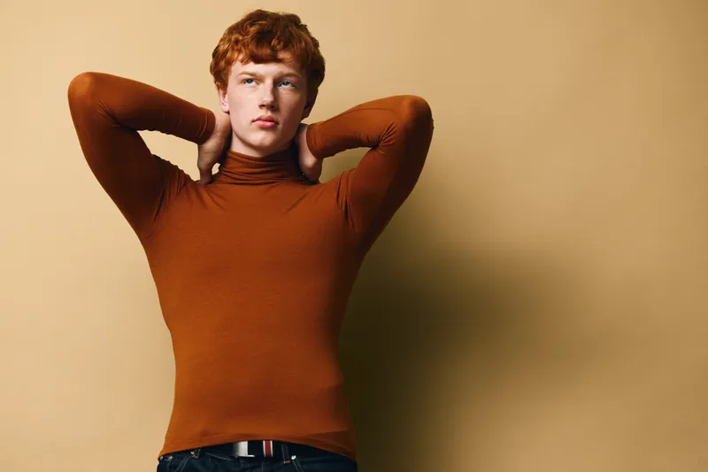 Young man with red hair wearing brown turtleneck posing with hands behind neck against beige background. Casual clothing, confident expression and studio portrait style.