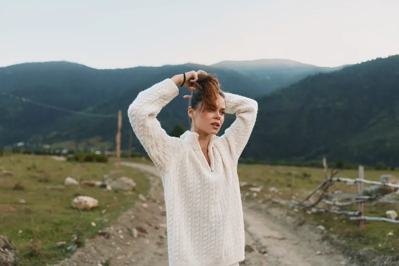 Young woman in a cozy white knit sweater stands outdoors in a mountain valley, adjusting her hair with a calm, contemplative expression under soft light, her posture relaxed, conveying solitude and