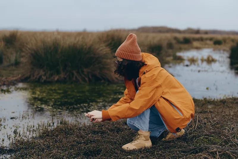 Woman lifestyle outdoors nature countryside wetland scene, she kneels in muddy grass, wearing orange jacket, knit beanie and boots, hood up, calm mood, autumn daylight in scenic field