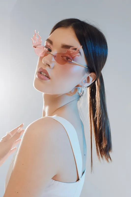 Young woman wearing butterfly-shaped pink sunglasses poses in a white tank top. Soft lighting highlights her beauty and fashion style in a minimalistic studio setting.