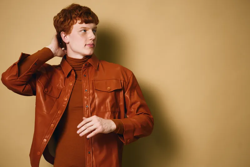 Young man with red hair wearing a brown jacket and turtleneck sweater standing against beige background looking sideways. Casual fashion, portrait and style with confident pose indoors.