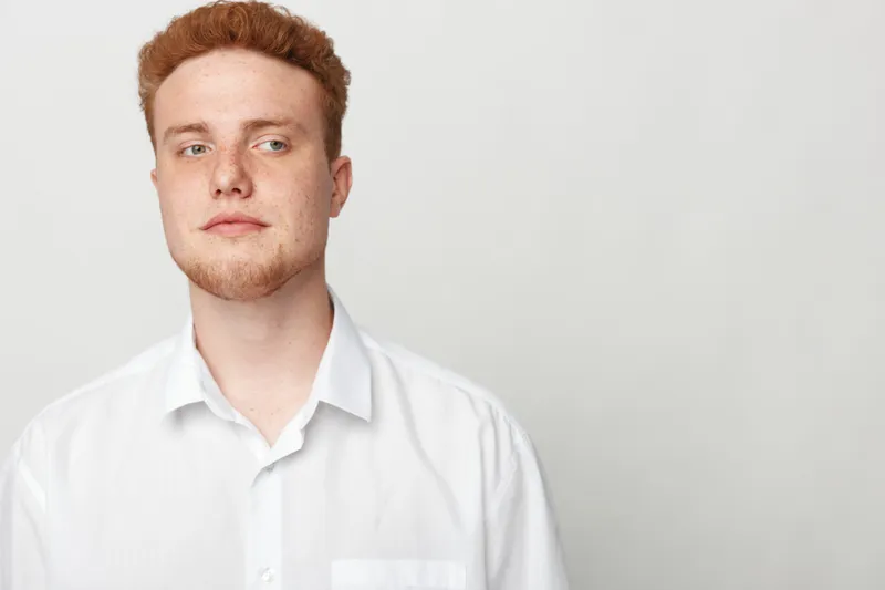 Young man with red hair and beard wearing white shirt looks confidently to the side on light background. Concept of casual style, youth, and natural portrait.