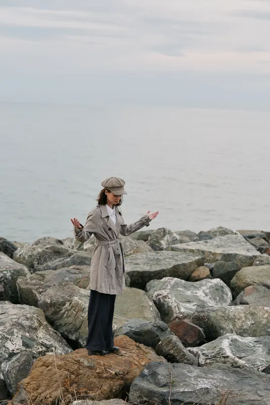 A woman in a patterned coat and hat stands on rocks by the calm sea, looking thoughtful. The cloudy sky and muted colors create a peaceful coastal scene.