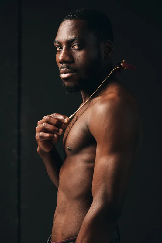 African man with dark skin and bare torso holding dried flower in creative conceptual visual style portrait with dramatic lighting and artistic expression in studio.
