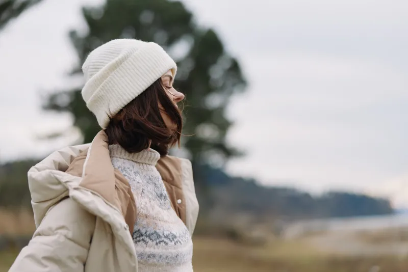 Winter woman in a cozy coat and beanie gazes into a wide outdoor landscape, soft light, chilly air, subtle motion and calm seasonal fashion.