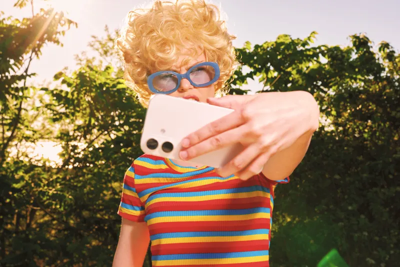 A woman with curly blond hair in a striped shirt selfies outdoors, blue sunglasses, retro film color vibe, capturing casual, everyday lifestyle moments in bright daylight.