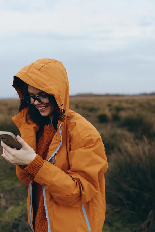 Woman smartphone outdoors raincoat hood lifestyle autumn smiling countryside nature, orange hooded raincoat, she walks in rural fields, looking at her phone with a relaxed smile
