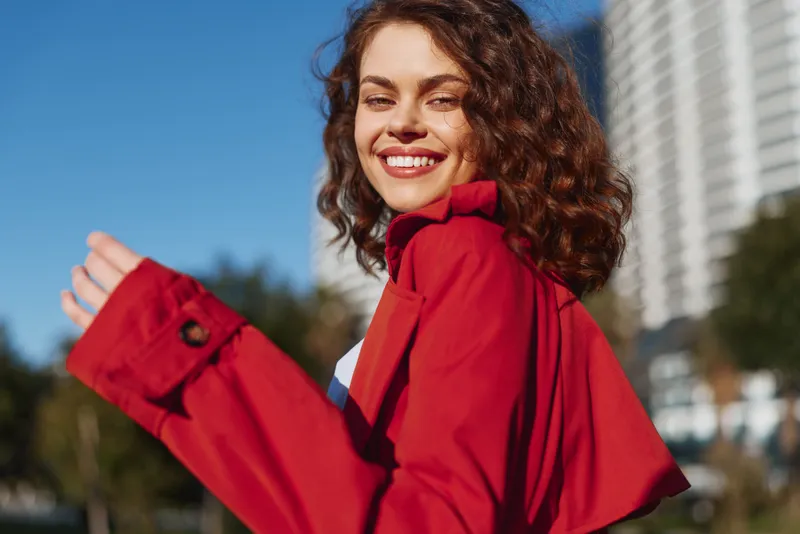 Smiling young woman with curly hair wearing red coat enjoys sunny day outdoors, looking back with joyful expression in urban environment with blurred buildings background.