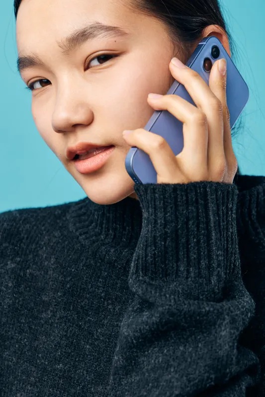 Young woman in black sweater talking on smartphone closely holding mobile device near ear with serious expression on blue background in studio shot portrait.