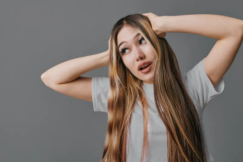 Young woman with long hair and surprised expression touching head on gray background. Casual style, emotional reaction with natural light and plain backdrop in studio.