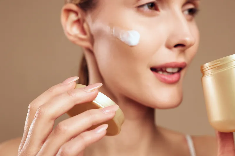 Woman applying face cream skincare wellness self care close up portrait cheek with fingers on brown background Soft studio lighting shows hydration and gentle skin treatment