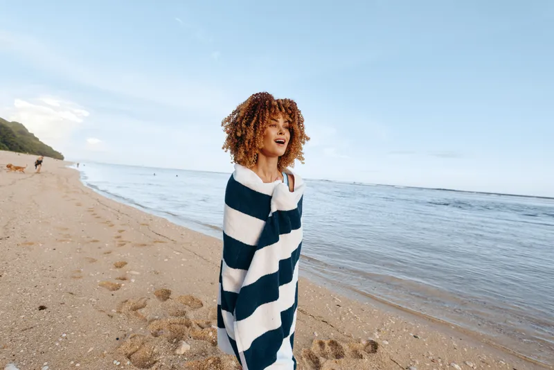 Young woman with curly hair wrapped in striped blanket stands on sandy beach near calm sea under clear sky enjoying peaceful moment.