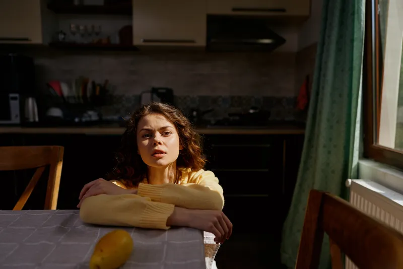 Woman leaning on a kitchen table, sunlit moment, casual home lifestyle, relaxed posture, candid expression, warm natural colors, table foreground, cozy kitchen background