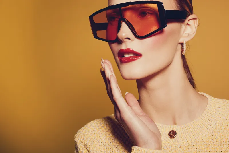 Stylish woman with oversized orange sunglasses and red lipstick poses against yellow background. Fashion concept with modern accessories and confident expression in studio lighting.