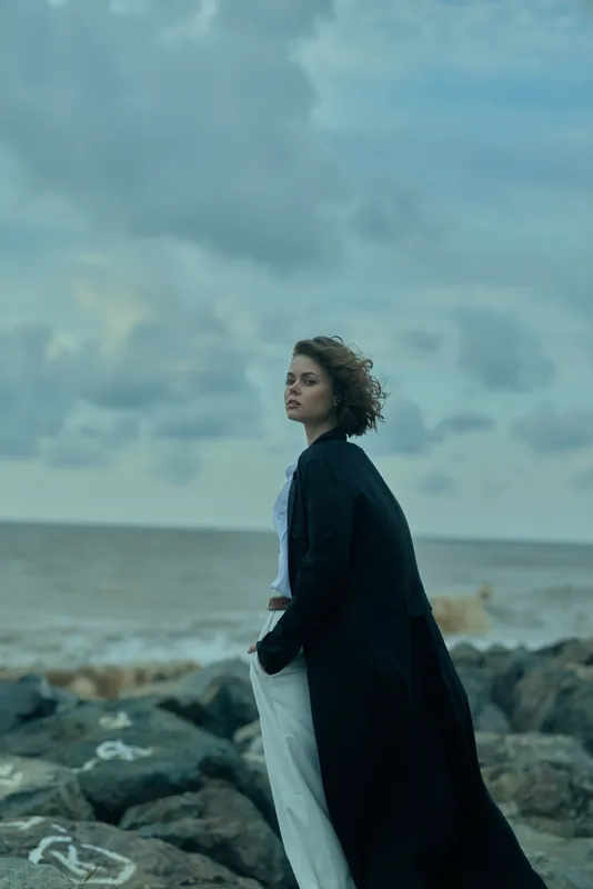 Woman in a long coat stands on a rocky shore as wind sweeps the scene, sea and horizon form a moody coastal seascape at dusk.