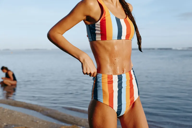 Young woman in a striped bikini posing on a sandy beach by the sea, summer sunlight, confident mood, soft waves in background, travel lifestyle, outdoors, close up, toned body, summer fun