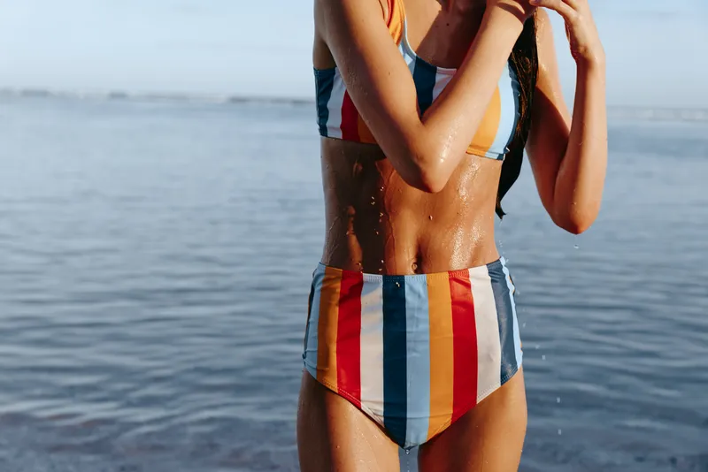 Woman, swimsuit, beach, ocean, summer, striped, tan, leisure, female model standing at shoreline, sunlit ocean waves behind, posing in swimwear, relaxed vacation mood, closeup