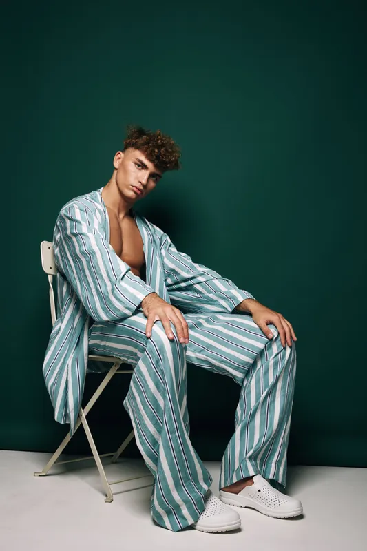 Young man sitting on chair wearing striped loose pajamas and white shoes against dark green background with relaxed pose and casual fashion style in studio setting.
