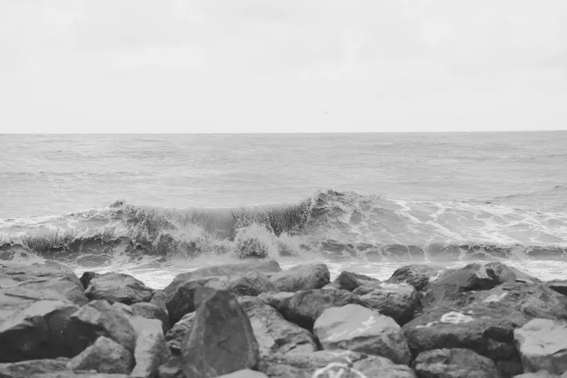 Rocks line the shore as gentle waves crash over stone, a calm seascape in grayscale capturing texture, weathered surfaces, and the quiet rhythm of the coast