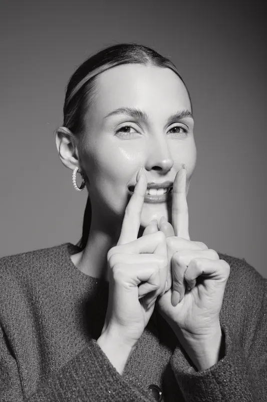 Smiling Caucasian woman with light skin wears hoop earrings and sweater, shows gesture with fingers near face. Studio portrait in black and white with soft lighting.