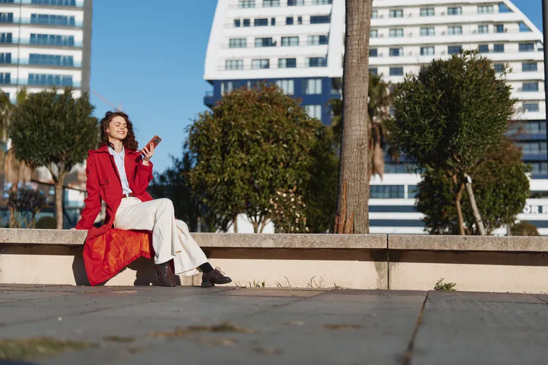 A stylish woman in a red coat sits on a concrete ledge in an urban park, checking her phone as bright sunlight highlights modern buildings.
