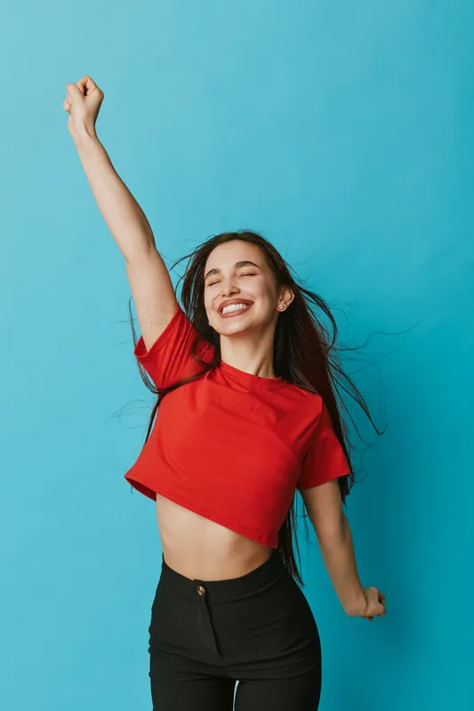 Young woman smiling and dancing with raised arm wearing red crop top and black pants on blue background. Happy girl with long hair expressing joy and freedom in studio.