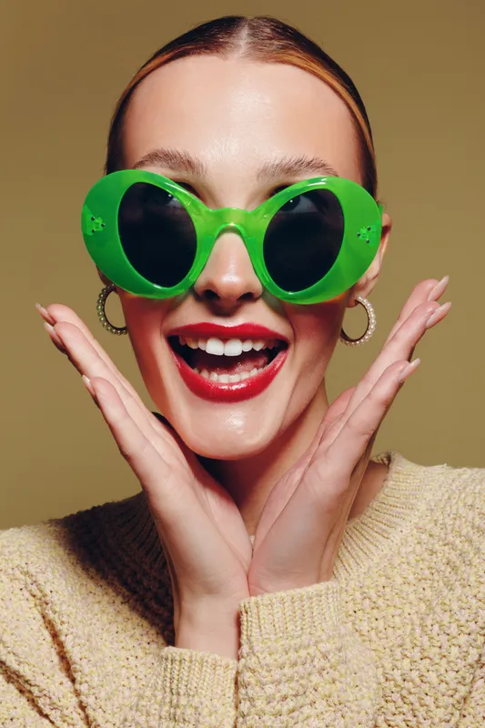 Happy young woman wearing large green sunglasses and red lipstick smiles widely with hands near face. Studio portrait with beige background and casual knitted sweater. Concept of joy and fashion.