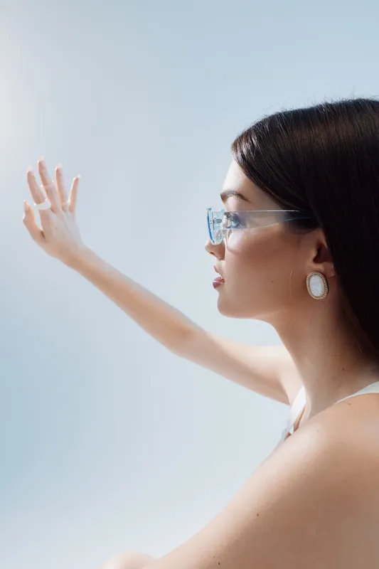 Profile of a young woman in clear glasses and elegant earrings reaching out with her hand against a light background. Concept of beauty, fashion, and modern style.