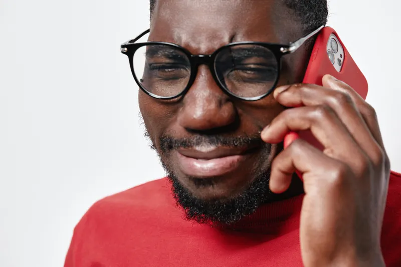 African American man with dark skin and glasses talking on red smartphone, wearing red shirt, expressing emotion with closed eyes on white background in studio portrait.