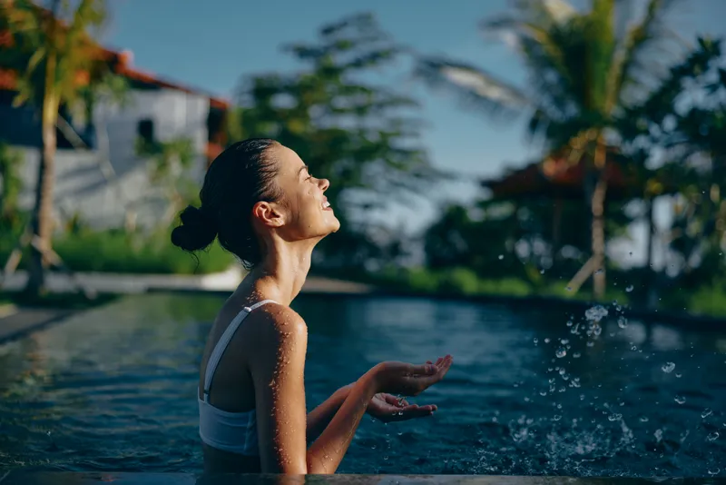 Young woman enjoys sunny day in outdoor swimming pool smiles with closed eyes splashes water with hands relaxed tropical palm trees background lifestyle concept