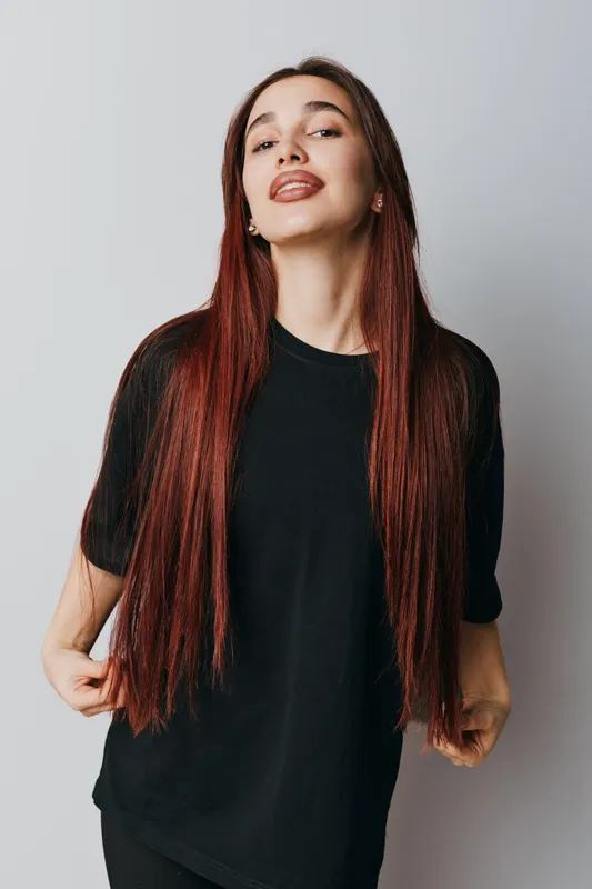 Young woman with long red hair wearing black t shirt standing against white background looking confident and smiling with hands holding hair in casual pose.