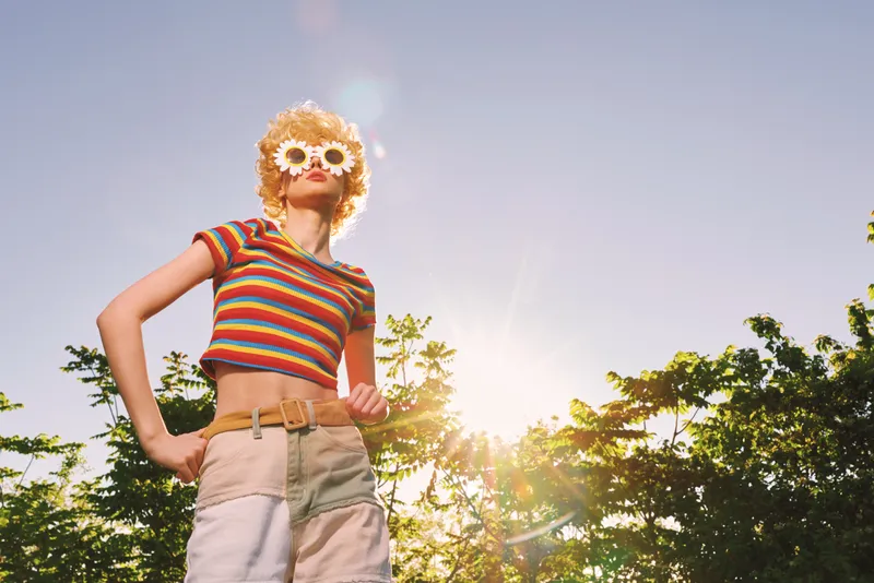 A young woman with curly blonde hair wearing a striped crop top and high waisted shorts poses outdoors in bright sun, retro color tones, sunglasses, radiating carefree confidence.