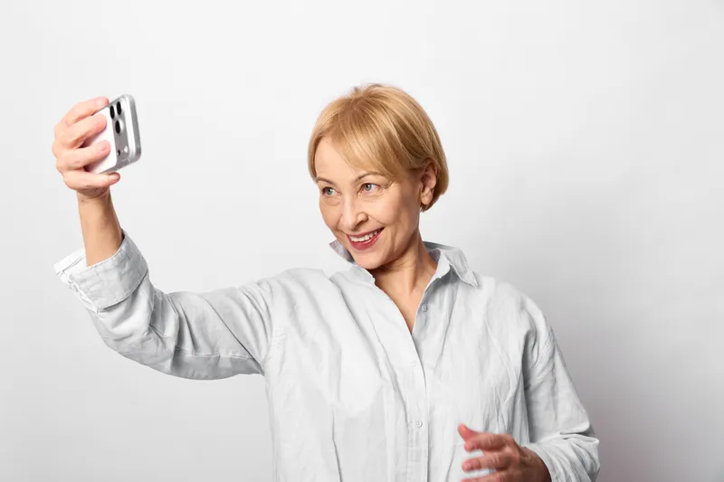 Older woman smiling for selfie, holding smartphone, portrait on white background, casual home clothes, friendly expression, studio light, copy space for social media, profile