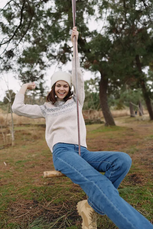 Young girl on a wooden swing in a park, smiling and relaxed, wearing a sweater and beanie, enjoying an outdoor moment among trees.