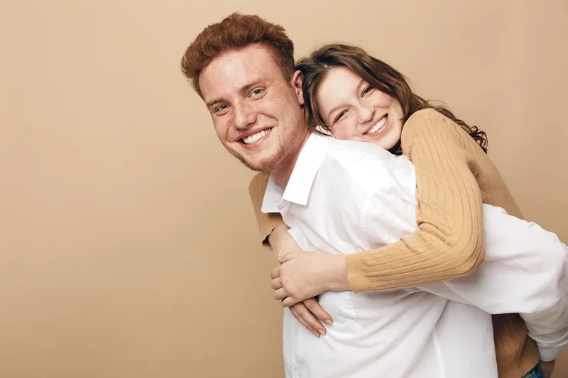 Happy young couple with red hair and brown hair smiling, woman hugging man from behind. Casual clothing and beige background. Concept of love, friendship, and joyful relationship.