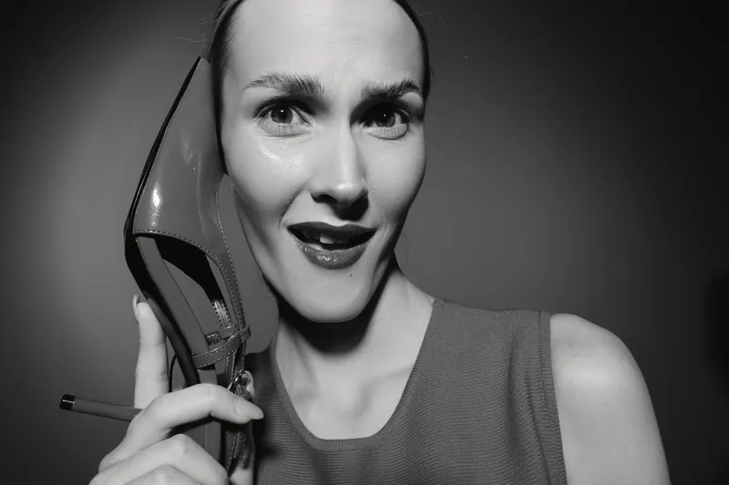 Young white woman with pale skin holds high heel shoe near face with surprised expression in black and white studio portrait.