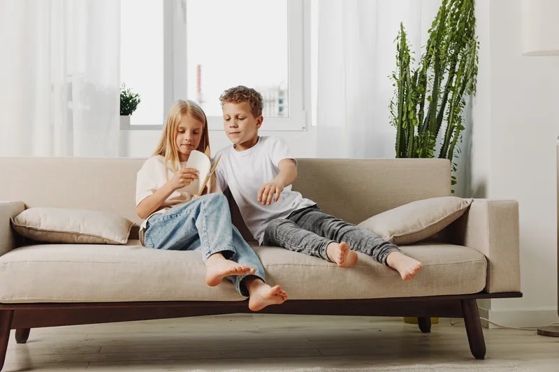 Two children sitting barefoot on beige sofa at home, looking at smartphone together in bright living room. Concept for family, technology, and childhood leisure time.