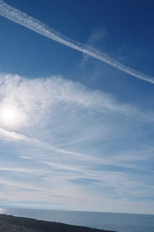 Bright sky over calm ocean with wispy clouds and contrails crossing above a quiet beach, capturing vast horizon, sea breeze and serene coastal atmosphere.