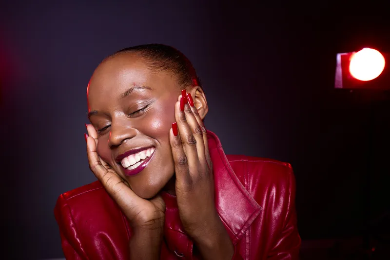 Smiling African American woman with dark skin and red painted nails enjoys a moment of happiness in a red leather jacket. Studio lighting highlights her glowing face and joyful expression.