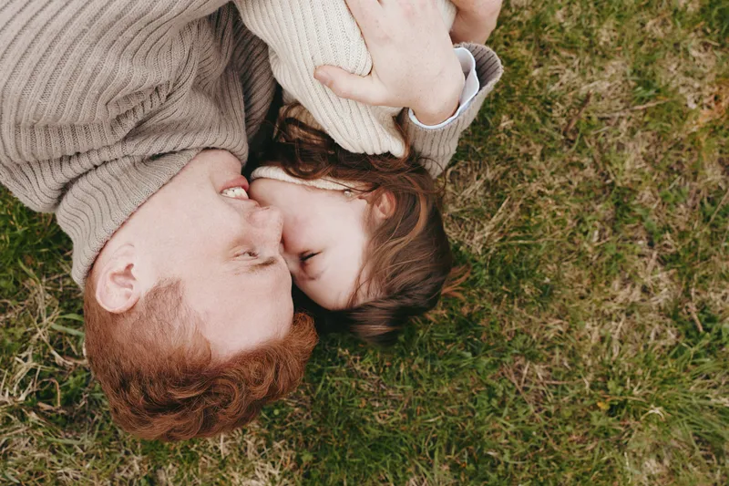 Redhead father and young daughter lie on grass embracing tenderly in a warm moment of family connection and love outdoors in casual sweaters during autumn.