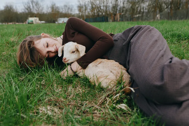 Young woman with long hair lies on the grass embracing a baby goat in a peaceful outdoor setting, capturing a tender moment of connection and relaxation in nature.
