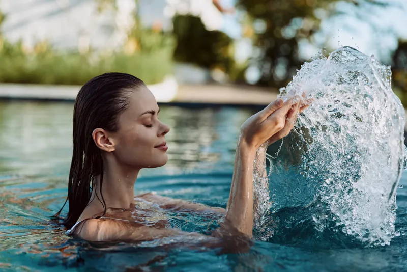 Woman enjoying leisure in a sunny pool, a splash as she plays with water, capturing relaxation and a carefree lifestyle beside clear blue water.