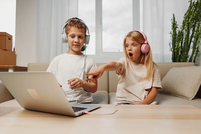 Two children with headphones sit on sofa using laptop, girl surprised by sound, boy focused on device. Home environment, casual clothing, technology, entertainment concept for kids.