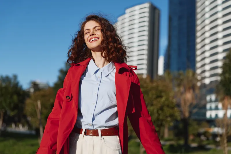 A smiling woman walks outdoors in a red coat and pale shirt, city high rises behind. Bright daylight emphasizes confident street fashion and carefree mood.