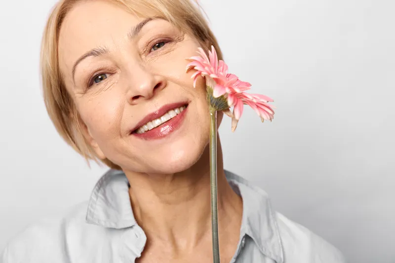 Woman smiling pink flower portrait closeup, mature woman with short hair. Bright studio lighting on white background creates warm calm wellness mood and gentle beauty concept.