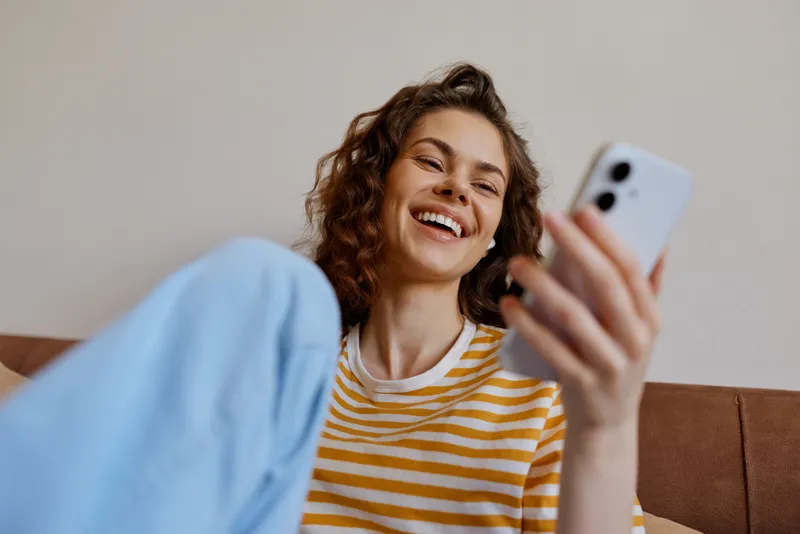 Young woman with curly hair lounges on a sofa at home, smiling as she scrolls through her phone, capturing a casual candid moment of everyday relaxation and connectivity.